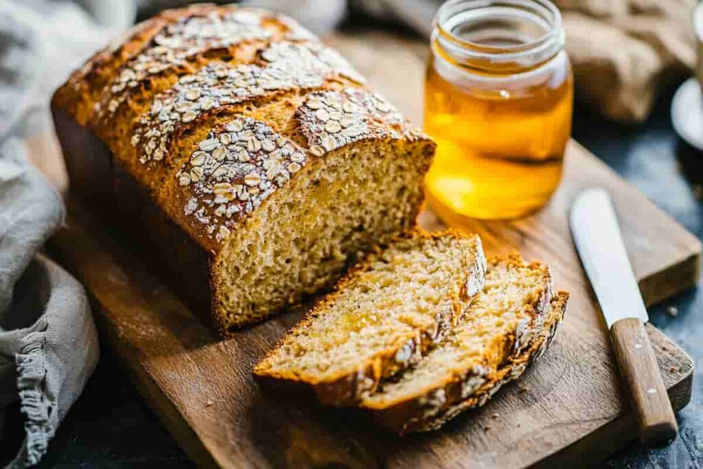 Sliced oat and honey bread loaf on a wooden cutting board, with a jar of honey and a butter knife nearby.